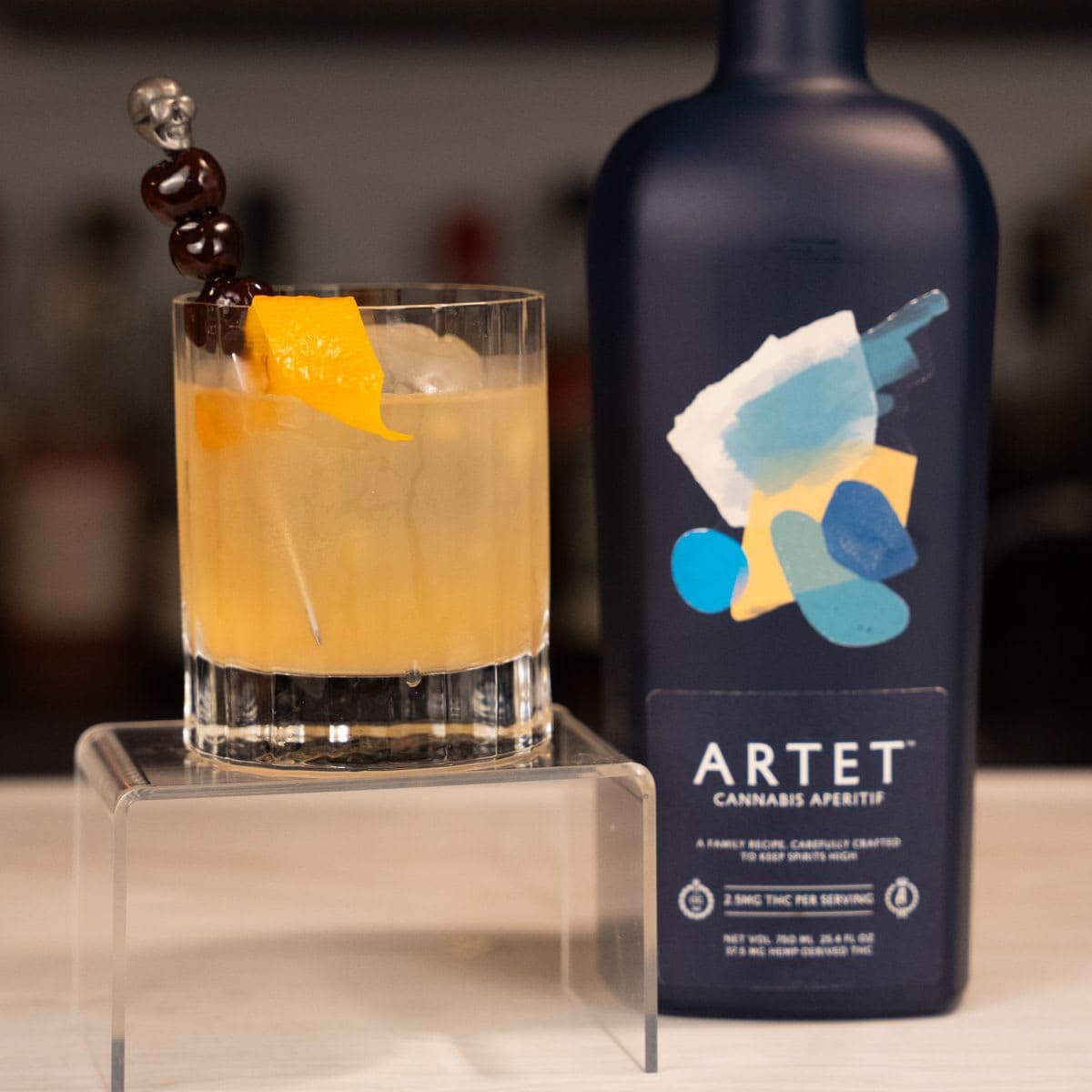 Cannabis smoked old fashioned with skull cherry pick and orange twist beside a bottle of Artet cannabis aperitif