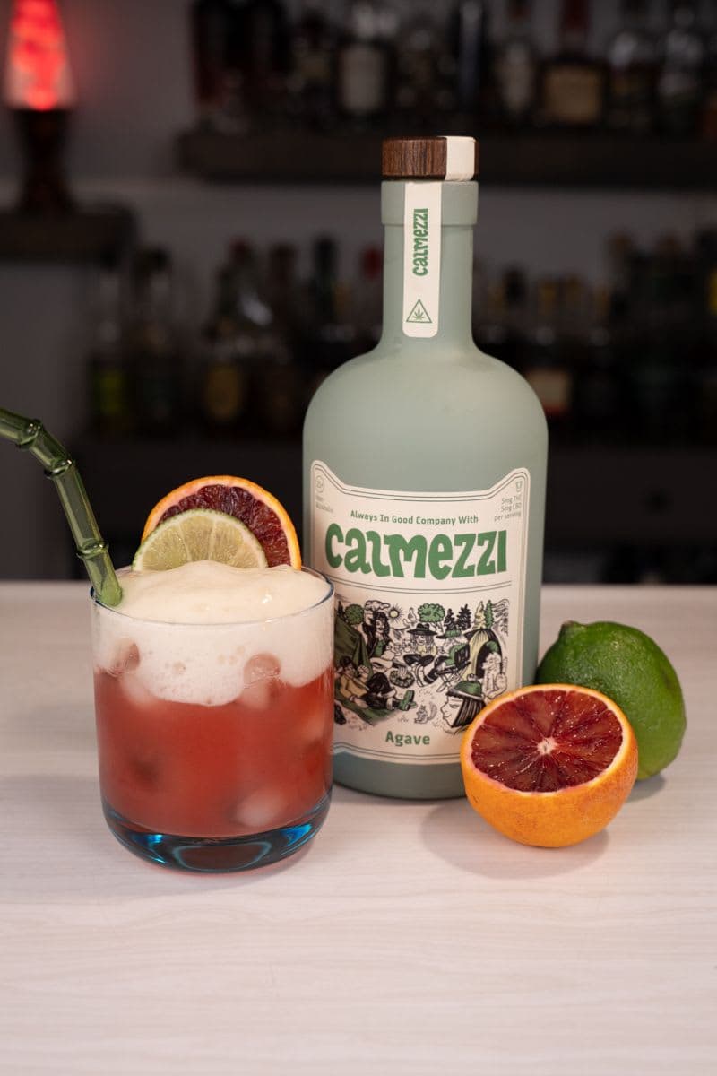 THC margarita topped with beer foam next to a bottle of Calmezzi agave THC spirit, blood orange and lime